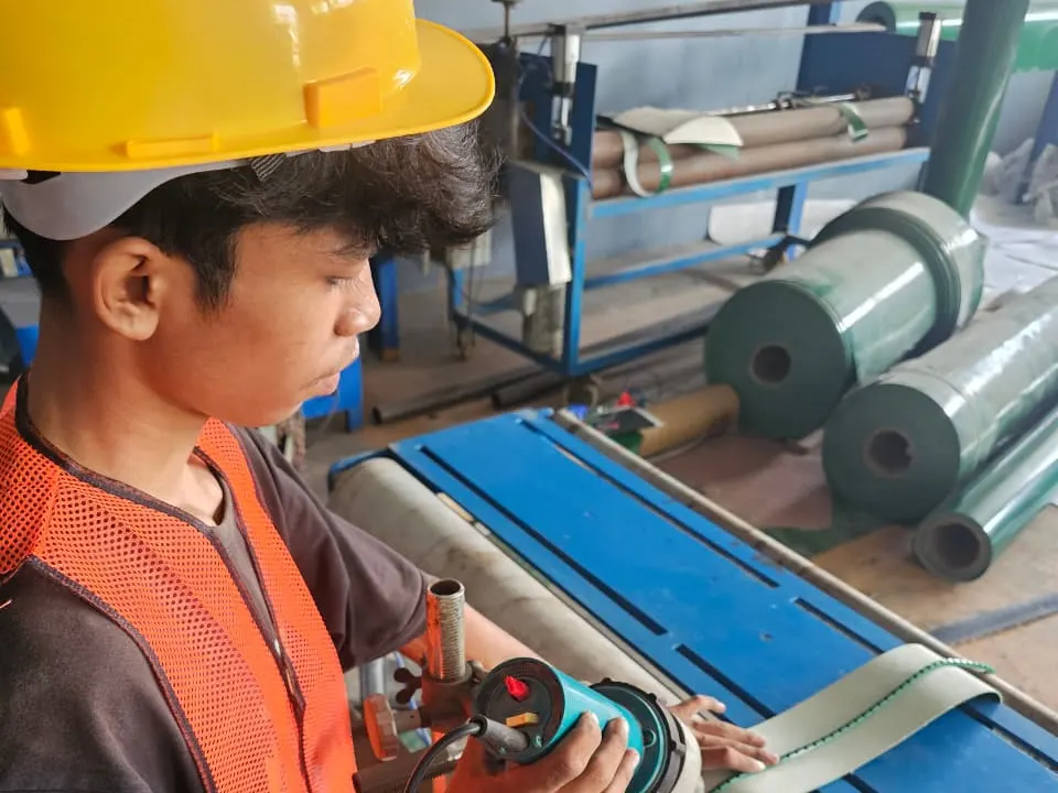 Teknisi BBS berhelm dan rompi hi-vis melas profile guide hijau ke punggung belt PVC putih menggunakan mesin las udara panas di workshop