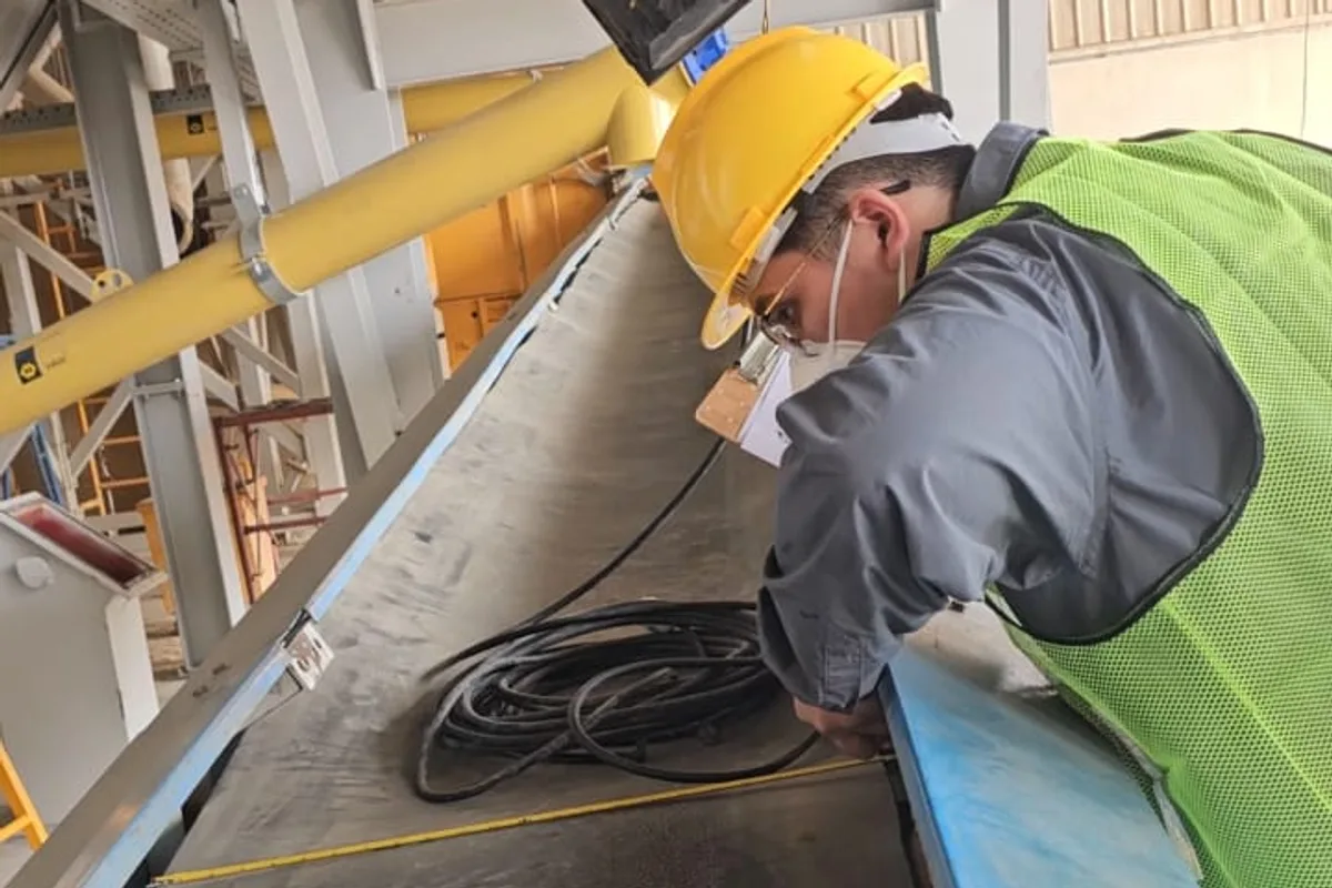 Teknisi berhelm dan rompi hi-vis mengukur belt conveyor dengan meteran langsung di jalur produksi pabrik pelanggan
