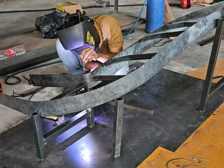 Teknisi BBS mengelas rangka conveyor melengkung dengan pelindung wajah di workshop, percikan busur las terlihat di sepanjang sambungan